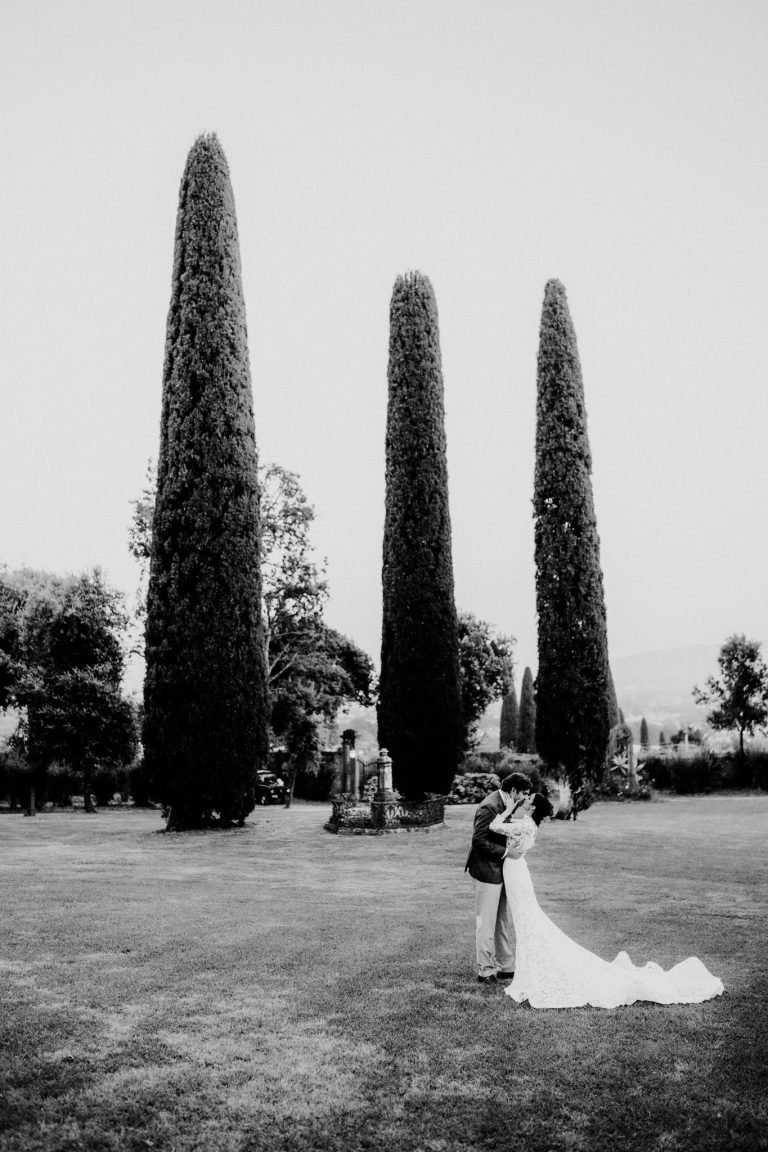Bride and groom sharing a romantic kiss in the gardens of Villa La Badiola in Tuscany, surrounded by cypress trees – black and white photo